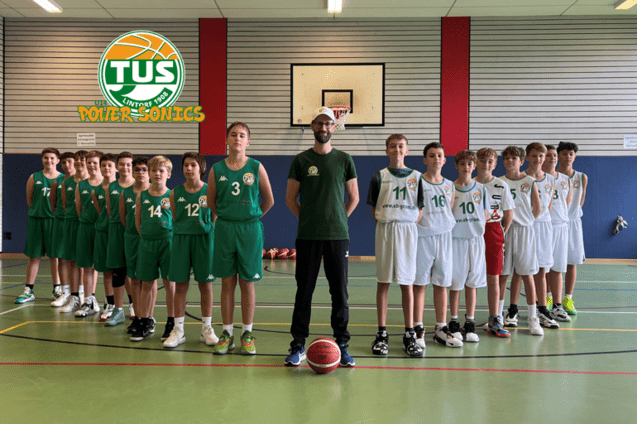 Gruppenshot von Basketballspielern in gr&uuml;nen und wei&szlig;en Trikots, aufgestellt in einer Sporthalle mit Basketballkorb.