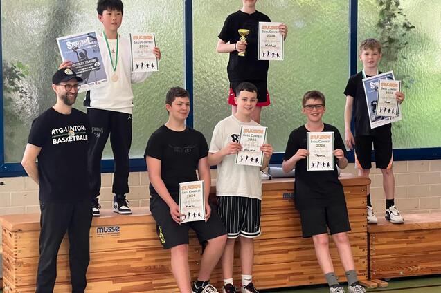 Gruppe von sechs Jugendlichen, die Pokale und Urkunden nach einem Basketballturnier in einer Sporthalle pr&auml;sentieren.