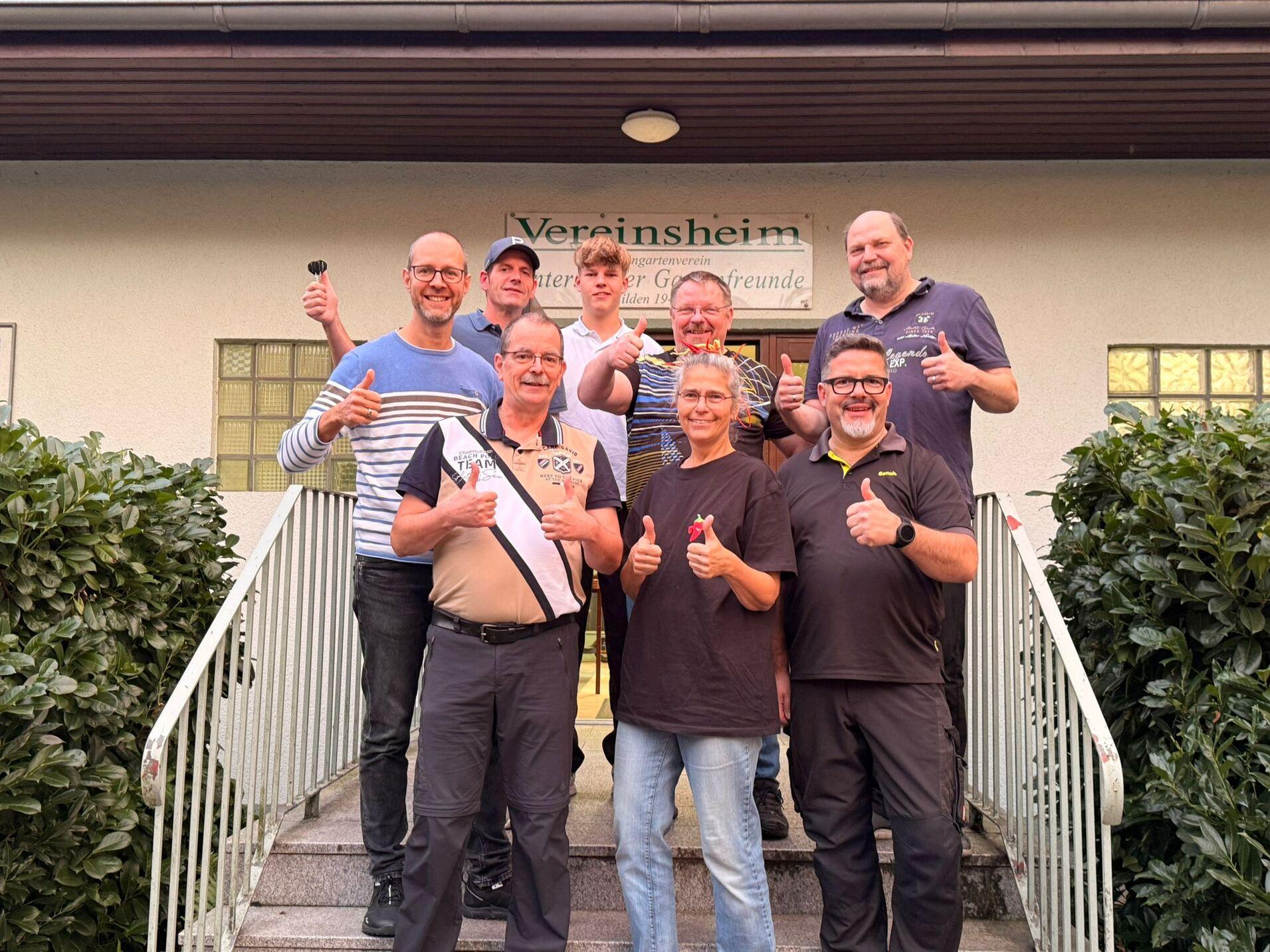 Gruppe von neun Personen vor einem Vereinsheim mit Daumen-hoch-Gesten und einer Urkunde in der Mitte.