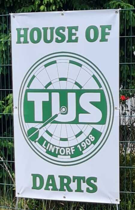 Banner mit dem Text „HOUSE OF TUS LINTORF 1908 DARTS“ auf einem Zaun, umgeben von grünem Laub.