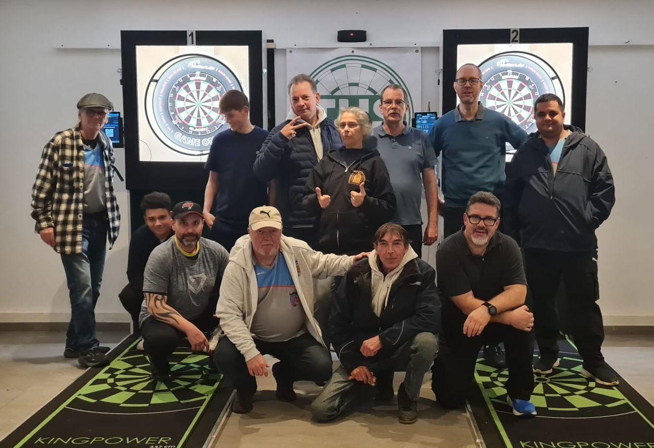 Gruppe von 12 Personen vor zwei Dartboards in einer Sporteinrichtung, mit Dartspiel-Ausrüstung und freundlich lächelnd.