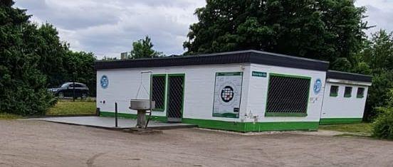 Weißes Gebäude mit grünen Akzenten, umgeben von Bäumen, im Vordergrund eine aufgeräumte Fläche.