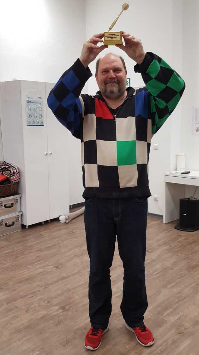 Mann mit buntem Pullover hält einen Pokal in die Luft, umgeben von einem Sporteinrichtungsraum.