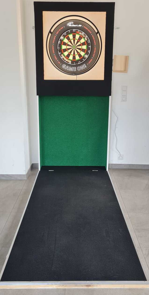 Dartboard an der Wand vor einer schwarzen Abwurfmatte in einem lichtdurchfluteten Raum.