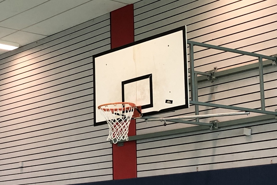 Korb und Brett eines Basketballst&auml;nders an einer Wand in einer Sporthalle mit grauen W&auml;nden.