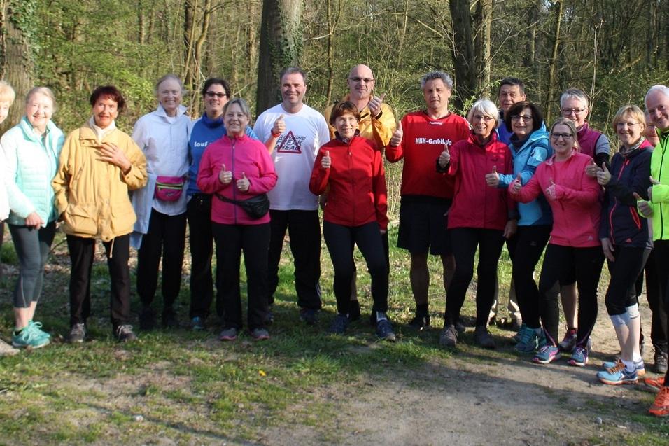 Gruppe von 15 Menschen in Sportkleidung steht auf einem Waldweg, umringt von B&auml;umen und auf gr&uuml;nem Gras.