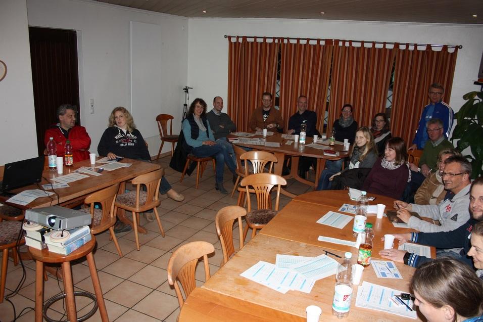 Gruppenbesprechung in einem Raum mit Tischen, St&uuml;hlen und Pr&auml;sentationstechnik; Teilnehmer h&ouml;ren aufmerksam zu.