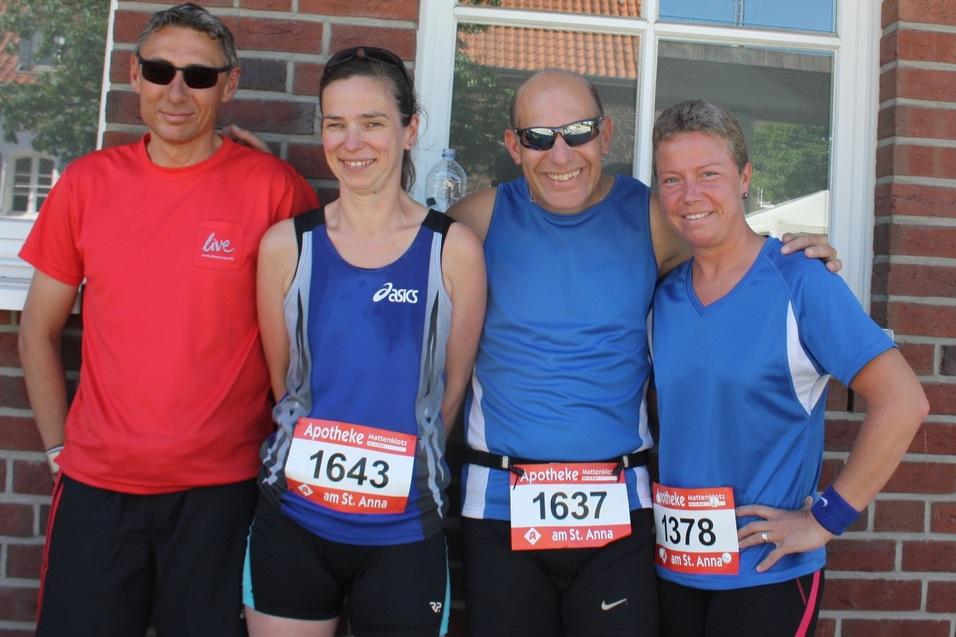 Vier Erwachsene in Sportbekleidung stehen l&auml;chelnd vor einer Backsteinmauer, mit Startnummern auf ihren Trikots.