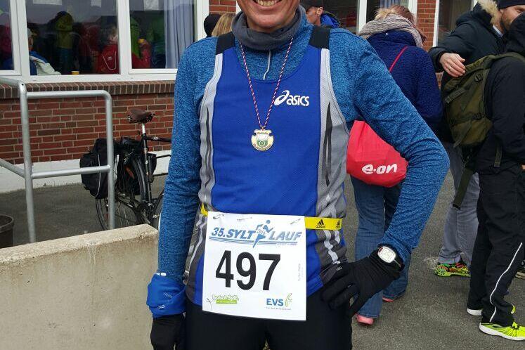 Lächelnder Läufer mit Medaille und Startnummer 497, im blauen Trikot vor einer Zuschauergruppe.