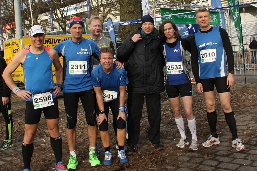 Gruppe von sechs Läufern in Sportkleidung, lächelnd vor einem Rennen, umgeben von Bäumen und Wettkampf-Elementen.