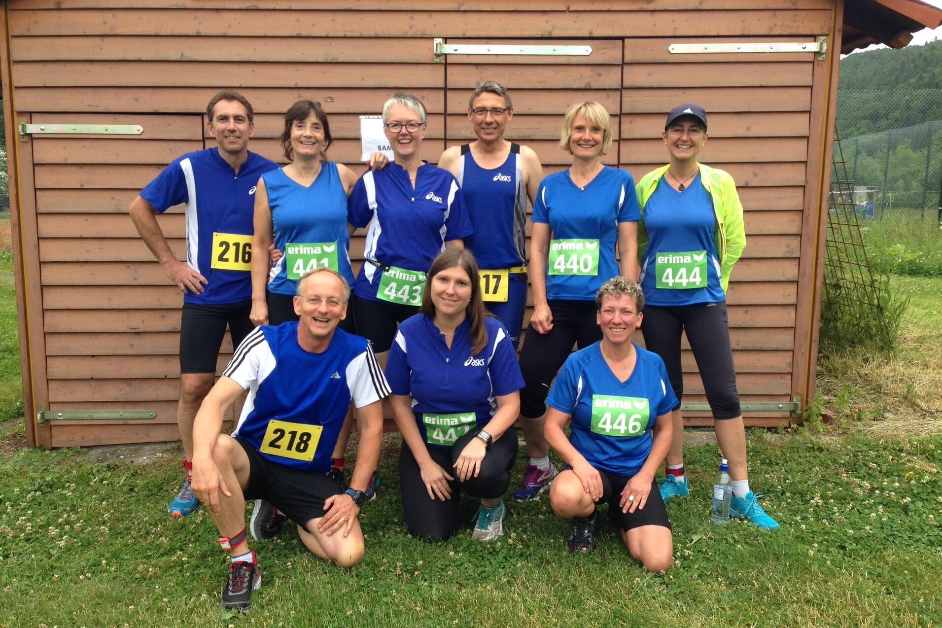 Gruppe von neun Läufern in blauen Trikots mit Startnummern vor der Angstberg-Arena, umgeben von grünem Gras.