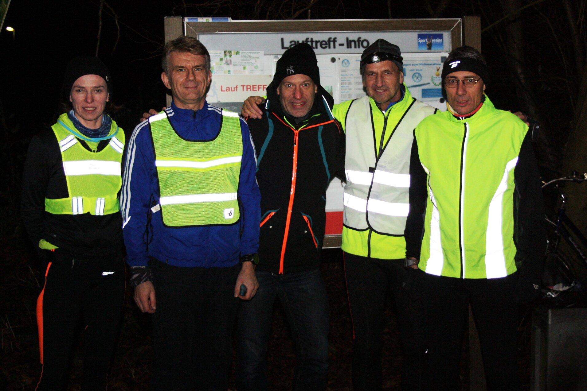 Fünf Personen in reflektierender Laufkleidung stehen vor einem Informationsschild für einen Lauftreff bei Nacht.