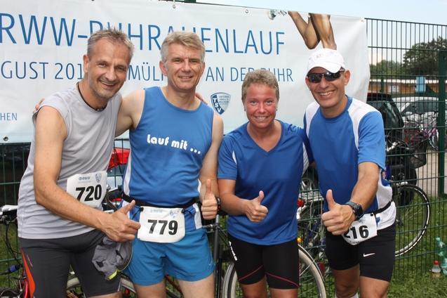 Vier Teilnehmer des RWW-Purhralenlaufs posieren l&auml;chelnd mit Daumen hoch vor einem Werbebanner.