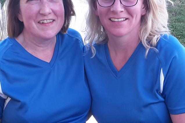 Zwei Frauen in blauer Sportkleidung l&auml;cheln, mit Startnummern auf den T-Shirts, im Hintergrund eine gr&uuml;ne Wiese.