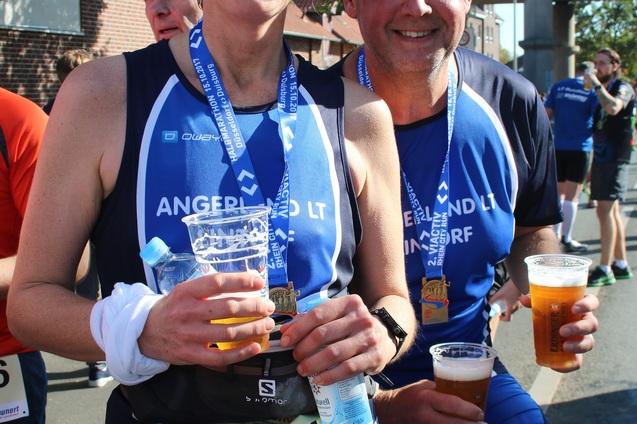 L&auml;chelnde L&auml;ufer mit Medaillen und Getr&auml;nken stehen nach einem Wettkampf nebeneinander. Sonntagswetter und Zuschauer im Hintergrund.