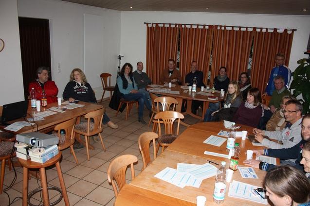 Gruppenbesprechung in einem Raum mit Tischen, St&uuml;hlen und Pr&auml;sentationstechnik; Teilnehmer h&ouml;ren aufmerksam zu.