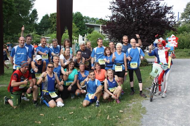 Gruppe von L&auml;ufern in blauen Shirts posiert l&auml;chelnd auf einer Wiese nach einem Wettkampf, begleitet von einem bunten Clown.