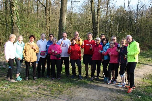 Gruppe von 15 Menschen in Sportkleidung steht auf einem Waldweg, umringt von B&auml;umen und auf gr&uuml;nem Gras.