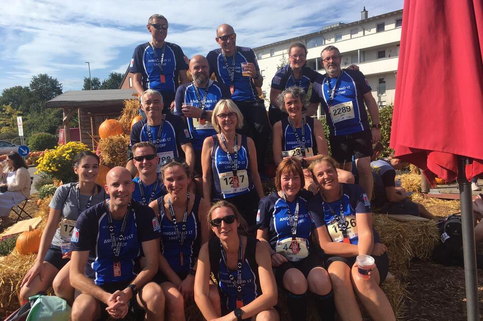 Gruppenzusammenkunft von Läufern in blauen Shirts mit Medaillen, sitzend auf Heuballen vor Kürbissen und einer Unterkunft.