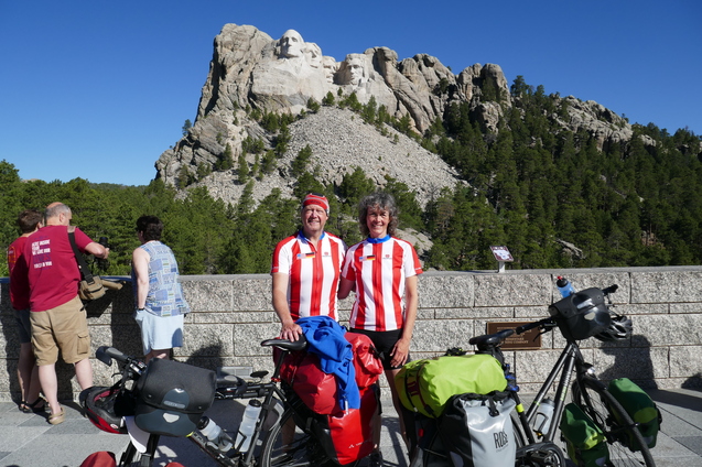 Zwei Radfahrer in rot-wei&szlig;en Shirts stehen vor dem Mount Rushmore, umgeben von B&auml;umen und Besuchern. Fahrr&auml;der sind sichtbar.
