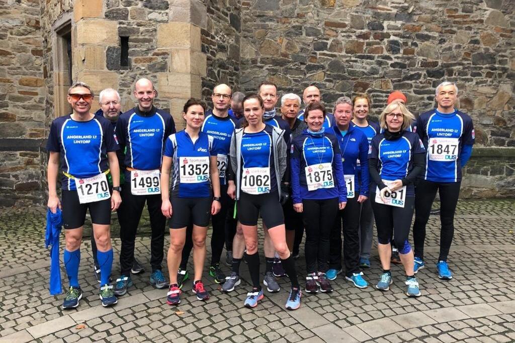 Gruppe von 16 Sportlern in blauen Trikots mit Startnummern, posierend auf einem gepflasterten Platz vor einer Steinmauer.