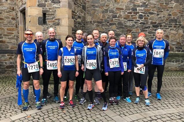 Gruppe von 16 Sportlern in blauen Trikots mit Startnummern, posierend auf einem gepflasterten Platz vor einer Steinmauer.
