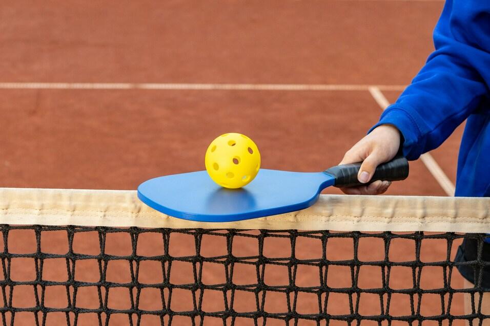 Nahaufnahme eines blauen Pickleball-Schlägers mit einem gelben Ball auf einem Netz von einem Tennisplatz.