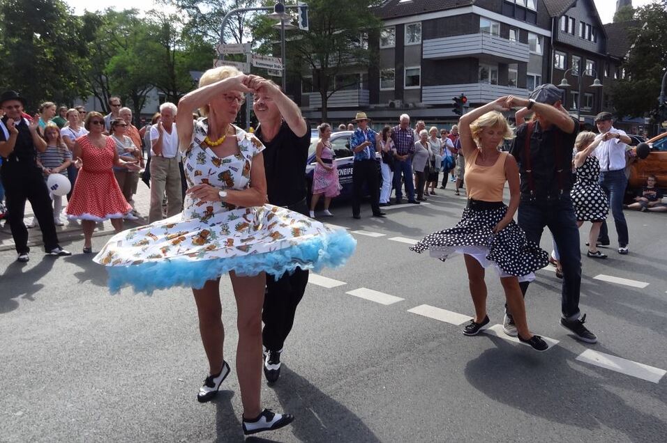 Tanzende Paare in bunten Kleidern auf einer Stra&szlig;e, umgeben von einer begeisterten Menge bei einer Veranstaltung.