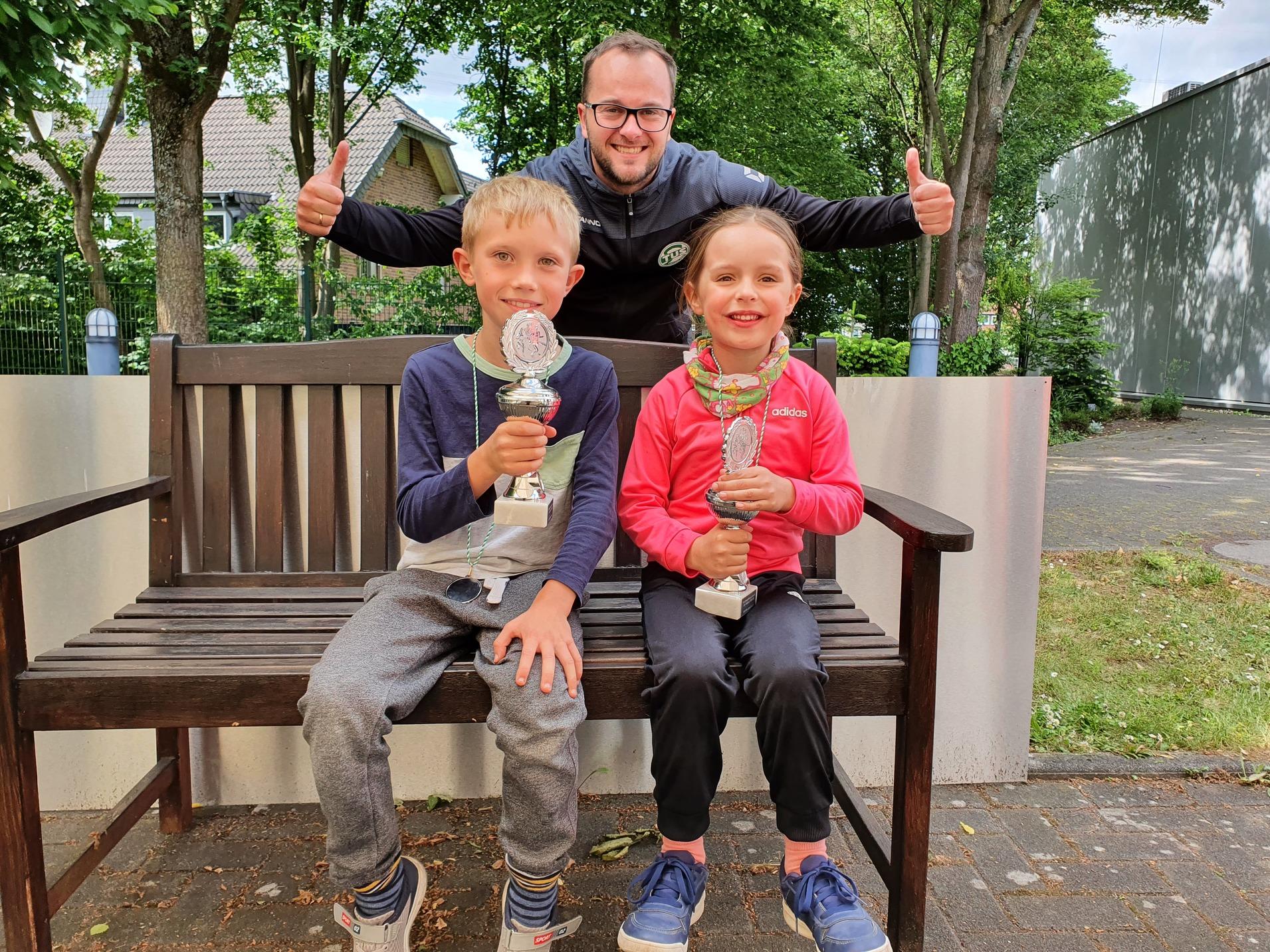 Zwei Kinder sitzen mit Pokalen auf einer Bank, während ein Erwachsener im Hintergrund jubelt.