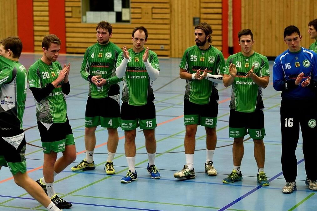 Sechs Handballspieler in gr&uuml;nen Trikots applaudieren in einer Sporthalle, w&auml;hrend ein siebter Spieler sich umdreht.
