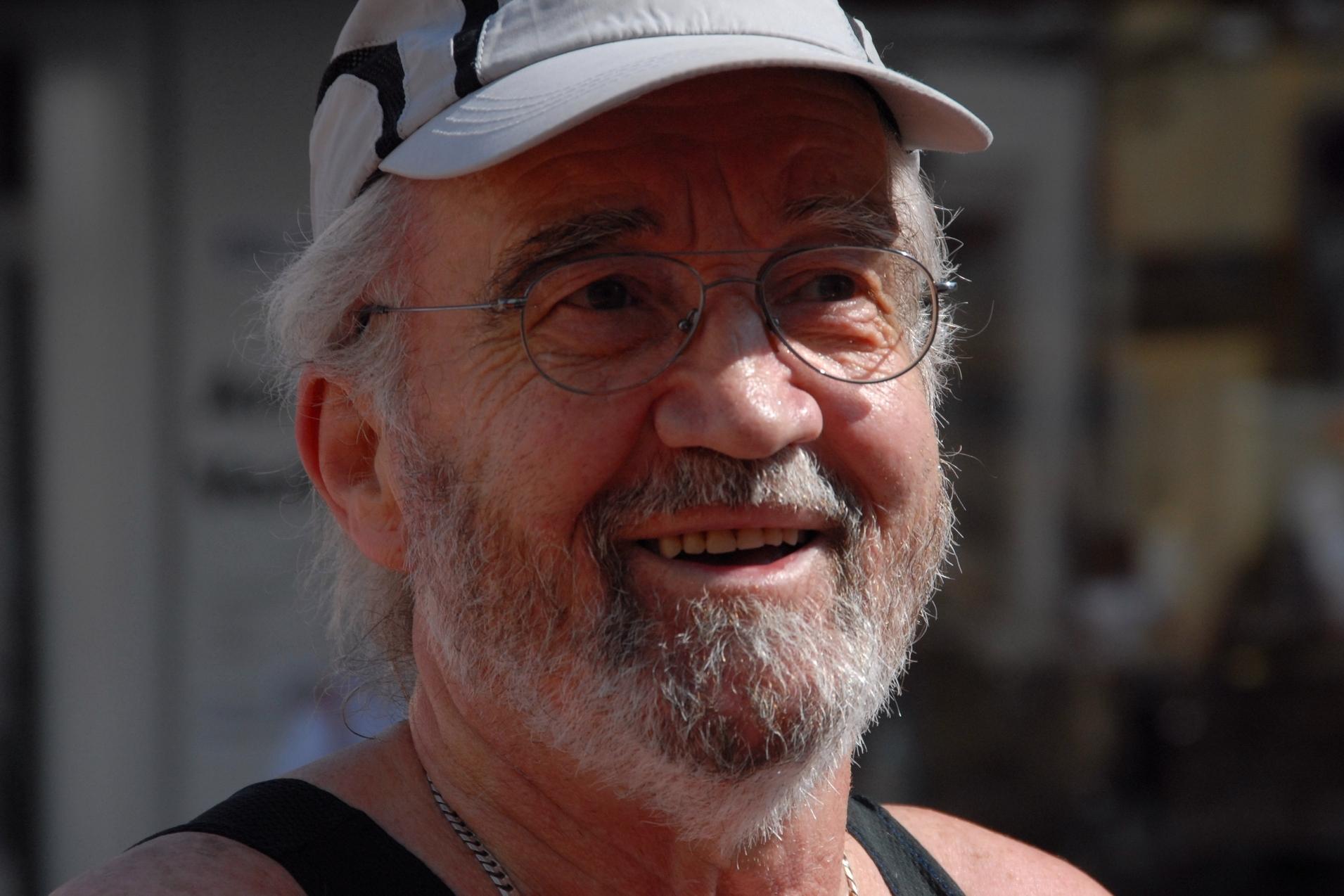 Lächelnder älterer Mann mit Brille und Schirmmütze, im Hintergrund verschwommene städtische Szenerie.