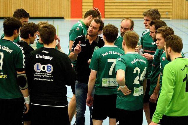 Eine Handballmannschaft bespricht sich, w&auml;hrend der Trainer Anweisungen gibt, in einer Sporthalle mit Holzw&auml;nden.