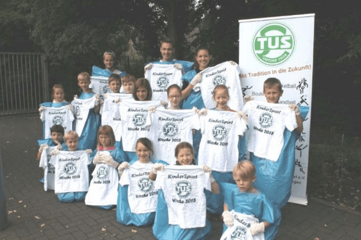 Gruppe von Kindern in Blauen Regenponchos, die T-Shirts mit der Aufschrift &bdquo;KinderSport Woche 2018&ldquo; halten.