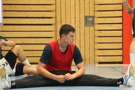 Junger Mann in rotem Trikot sitzt im Spagat auf einer Sporthalle, w&auml;hrend andere Spieler im Hintergrund sitzen.