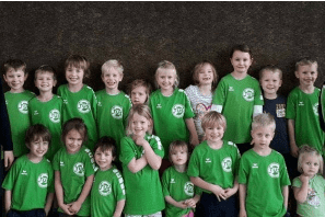 Gruppe von Kindern und Betreuern in gr&uuml;nen T-Shirts, l&auml;chelnd vor einem dunklen Hintergrund.