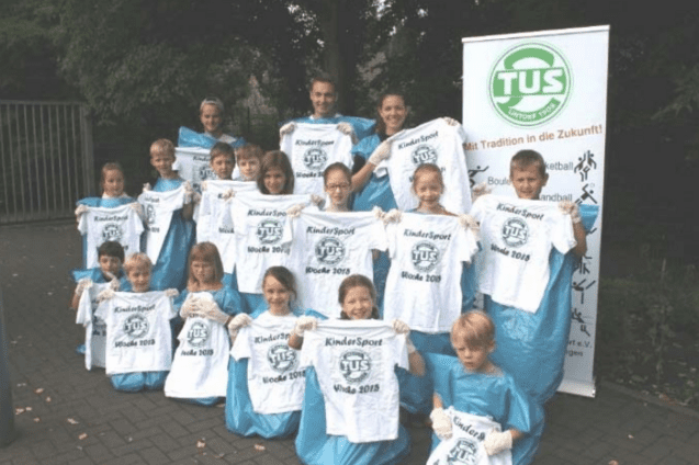 Gruppe von Kindern in Blauen Regenponchos, die T-Shirts mit der Aufschrift &bdquo;KinderSport Woche 2018&ldquo; halten.
