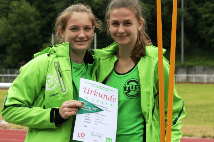 Zwei junge Sportlerinnen in gr&uuml;ner Sportkleidung halten stolz eine Urkunde auf einem Sportplatz.