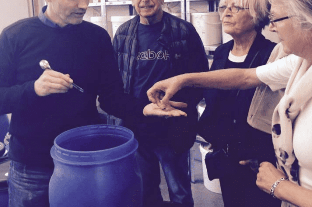 F&uuml;nf Personen stehen um einen blauen Beh&auml;lter und beobachten, wie ein Mann mit einem L&ouml;ffel etwas in eine Hand gibt.