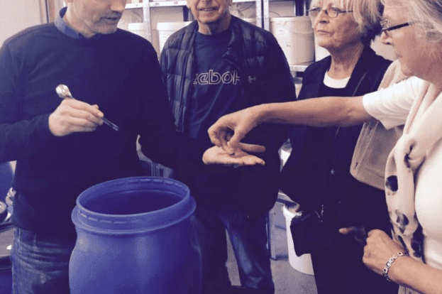 F&uuml;nf Personen stehen um einen blauen Beh&auml;lter und beobachten, wie ein Mann mit einem L&ouml;ffel etwas in eine Hand gibt.