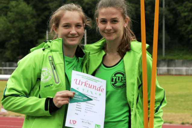 Zwei junge Sportlerinnen in gr&uuml;ner Sportkleidung halten stolz eine Urkunde auf einem Sportplatz.