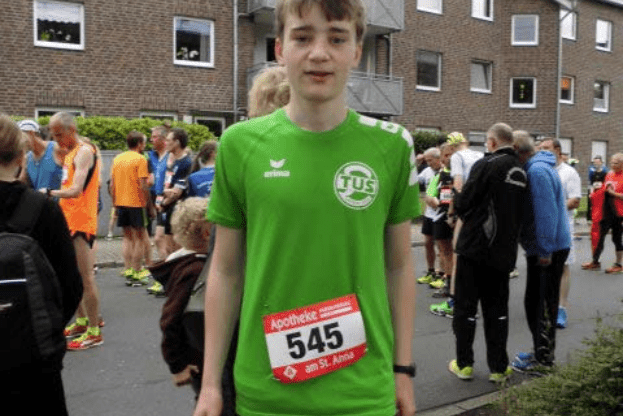 Junger L&auml;ufer mit gr&uuml;nem Trikot und Startnummer 545 steht vor einer Gruppe von Teilnehmern bei einem Laufereignis.