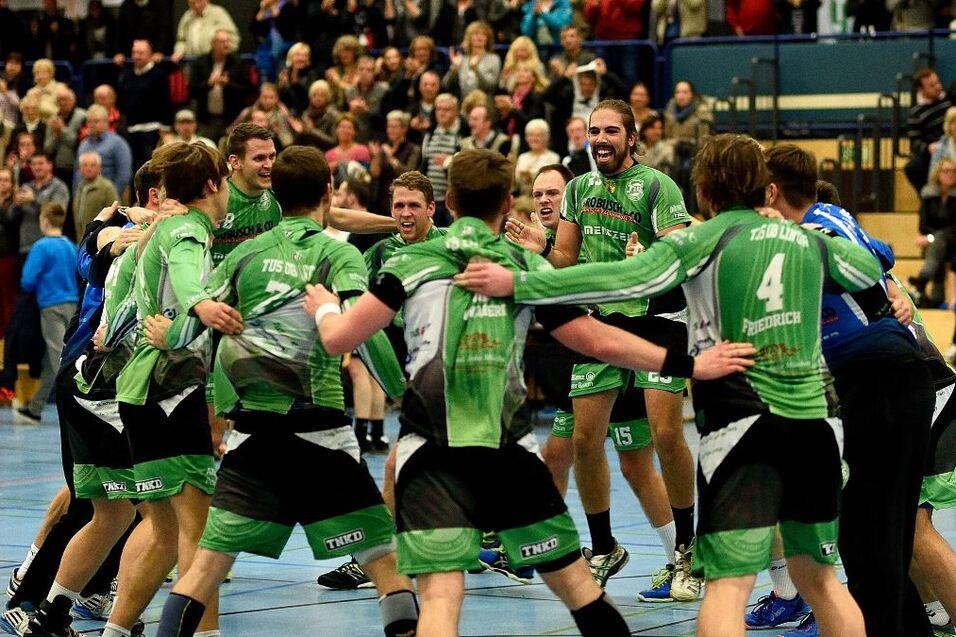 Feiernde Handballmannschaft in gr&uuml;nen Trikots umarmt sich in einer sportlichen Halle vor jubelndem Publikum.
