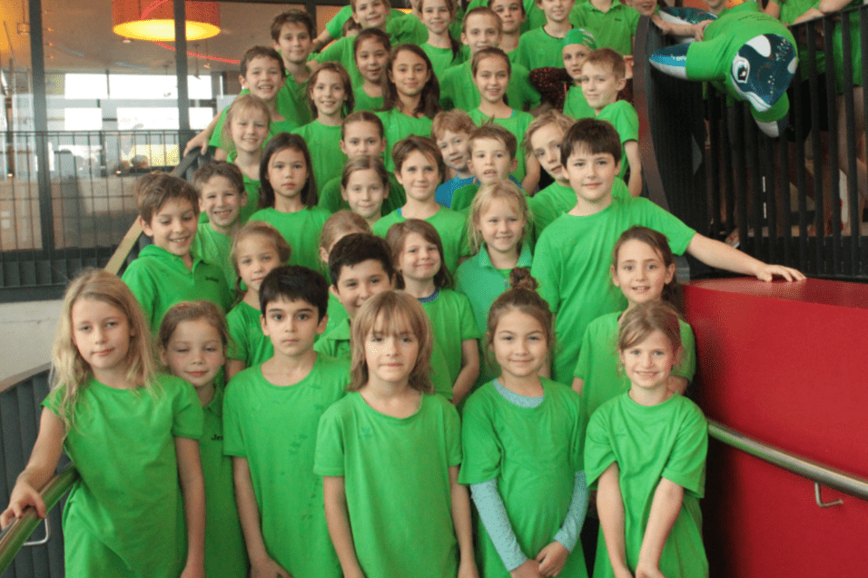 Gruppe von Kindern in gr&uuml;nen T-Shirts, die auf einer Treppe posieren, mit einem gro&szlig;en, gr&uuml;nen Luftballon im Hintergrund.