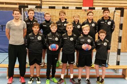 Gruppe von zw&ouml;lf Jungen in Sportkleidung, posierend vor einem Handballtor in einer Sporthalle.