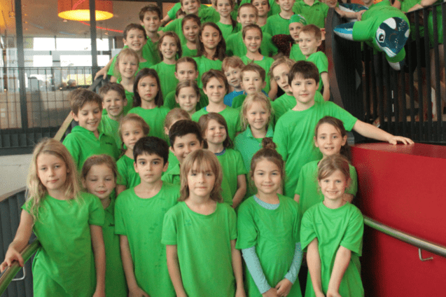 Gruppe von Kindern in gr&uuml;nen T-Shirts, die auf einer Treppe posieren, mit einem gro&szlig;en, gr&uuml;nen Luftballon im Hintergrund.