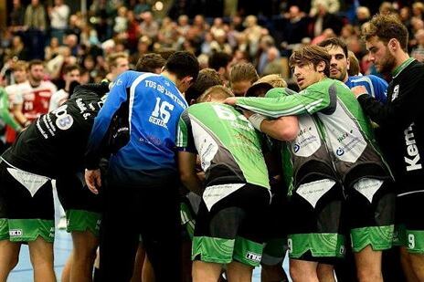 Handballmannschaft in einem Huddle, umgeben von Zuschauern in einer Sporthalle w&auml;hrend eines Spiels.