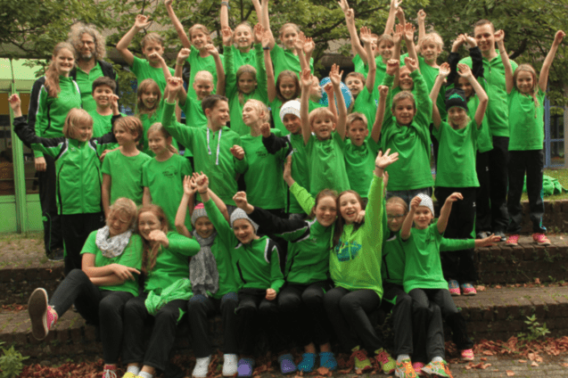 Gruppenschnappschuss von 40 Kindern in gr&uuml;nen T-Shirts, die fr&ouml;hlich auf einer Treppe posieren und die Arme heben.