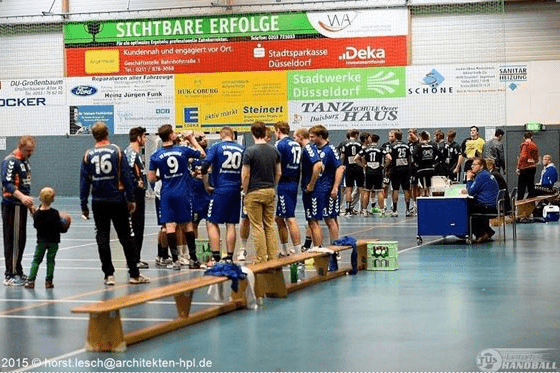 Handballspiel: Spieler in blauen Trikots stehen an der Bank, Zuschauer und Spieler warten auf das n&auml;chste Spiel.