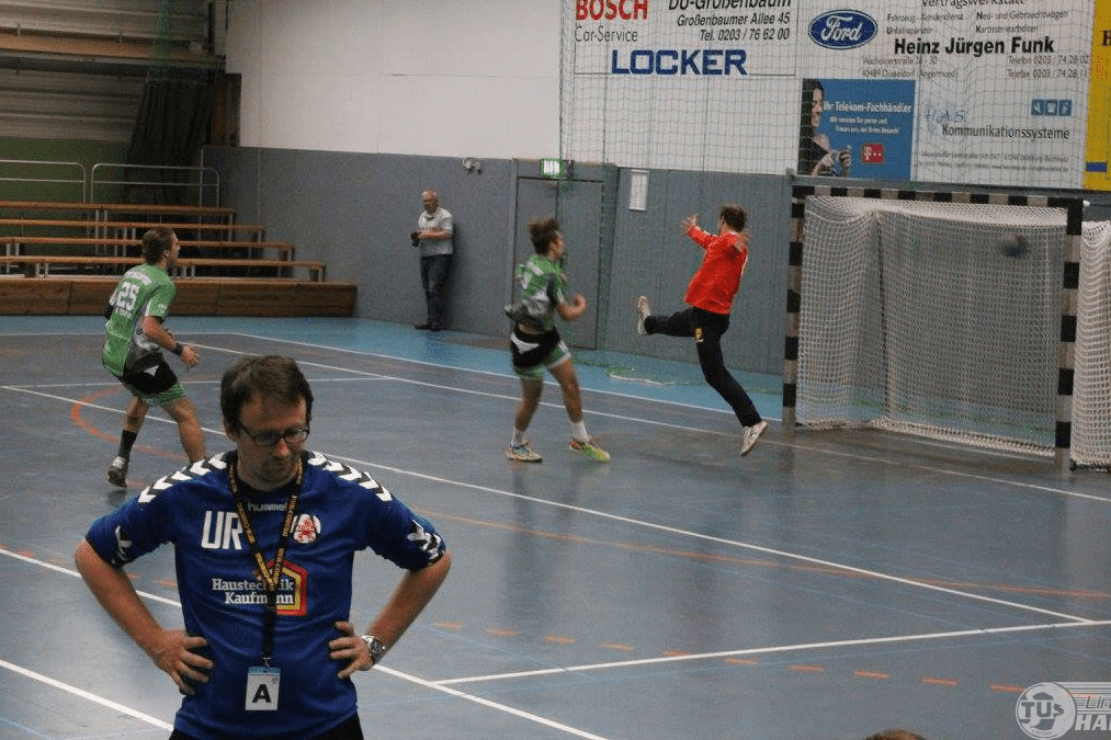 Handballspiel in der Halle: Spieler schie&szlig;en auf das Tor, w&auml;hrend der Torwart bereit ist. Ein Schiedsrichter &uuml;berwacht das Geschehen.