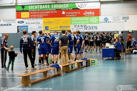 Handballspiel: Spieler in blauen Trikots stehen an der Bank, Zuschauer und Spieler warten auf das n&auml;chste Spiel.